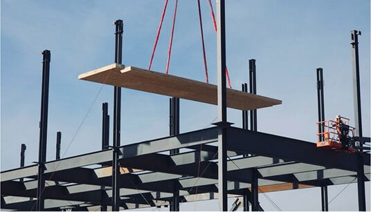 Crane lifting large wooden beam with red straps above steel framework structure at construction site under clear blue sky.