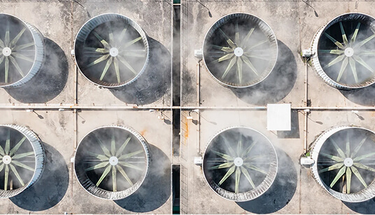 Aerial view of eight large industrial cooling fans arranged in two rows on concrete rooftop with mist rising.