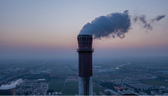 Tall smokestack emitting thick dark smoke above urban and agricultural landscape under hazy sky at sunrise or sunset.